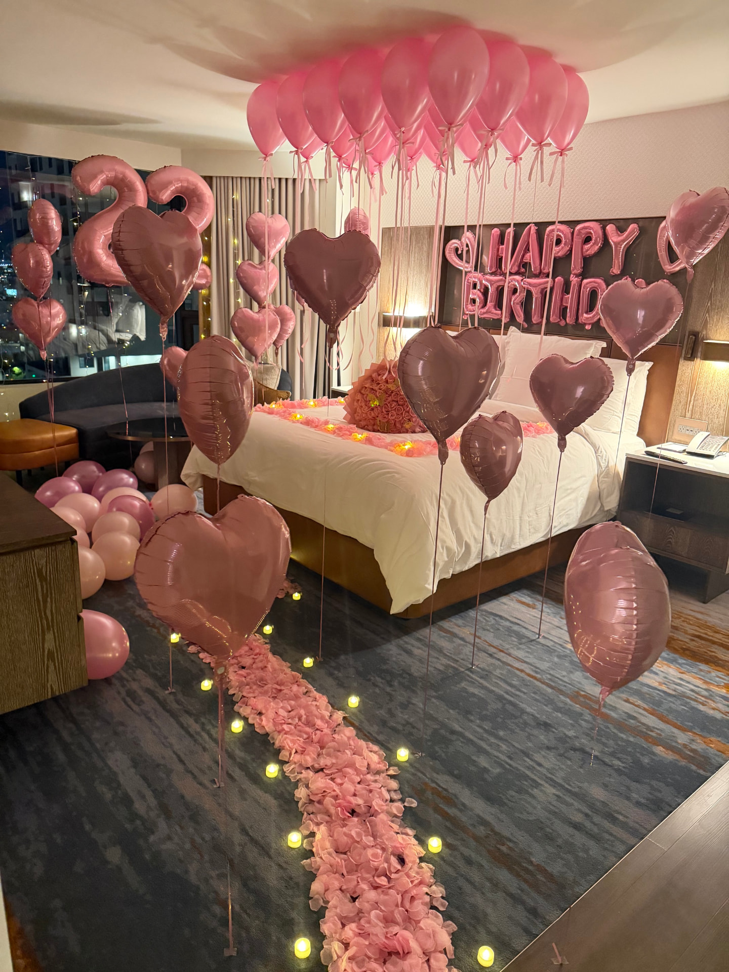 Hotel suite styled with a pink balloon ceiling, a Happy 21st Birthday balloon banner, heart balloons, and rose petals on the bed